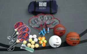 "Pickleball paddles, tennis rackets, basketballs, racket sport balls, a volleyball, and a Sport Court bag on a game court surface"