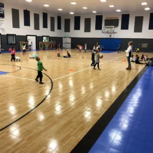 Groups of young kids in an indoor school gymnasium featuring Maple Select Flooring and a wall-mounted basketball hoop