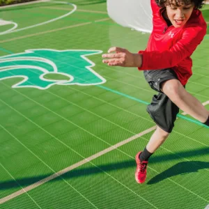A kid in a red and black uniform kicking on a green outdoor court surface