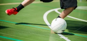 A foot with black socks and red shoes kicking a futsal ball on a green court surface