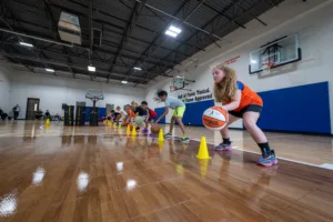 Kids lined up in a school gymnasium practicing basketball dribbling drills